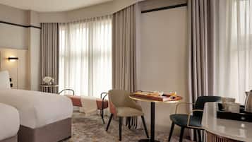Habitación de hotel con una cama grande, una mesa de comedor con desayuno, dos sillas, una mesita auxiliar con un jarrón y un ventanal con cortinas.
