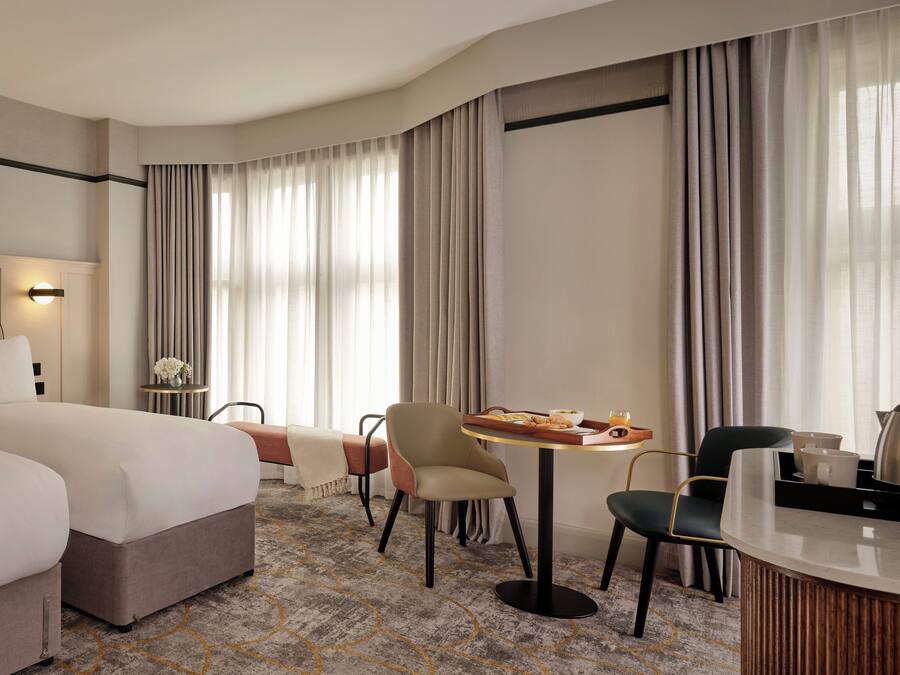 Quarto de hotel com uma cama grande, uma mesa de jantar com café da manhã, duas cadeiras, uma pequena mesa lateral com um vaso e uma janela ampla com cortinas.