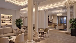 Un hall d’entrée spacieux avec de hauts plafonds, des colonnes richement décorées et un lustre. On y trouve des coins salon avec des canapés et des fauteuils, ainsi qu’un comptoir de réception à l’arrière-plan.