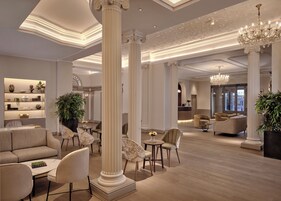 A spacious lobby with high ceilings, ornate columns, and a chandelier. There are seating areas with sofas and chairs, and a reception desk in the background.