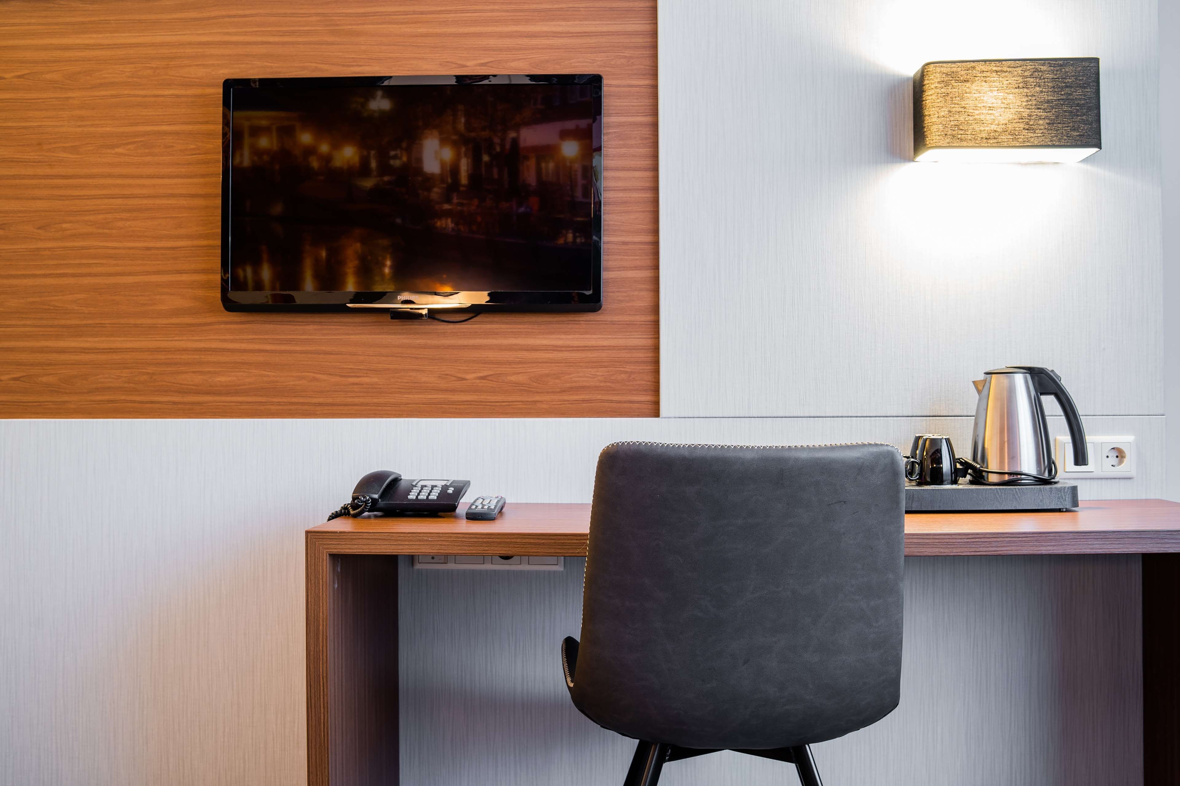A hotel room with a flat-screen TV mounted on a wooden wall, a desk with a telephone and remote controls, a black office chair, and a wall-mounted lamp.