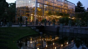 Un edificio moderno de cristal con fachada reflectante, rodeado por una pequeña fuente de agua y follaje.