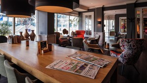 Un moderno espacio de bar con mostrador de madera, periódicos y diversas opciones de asientos, como sillas y un sofá.