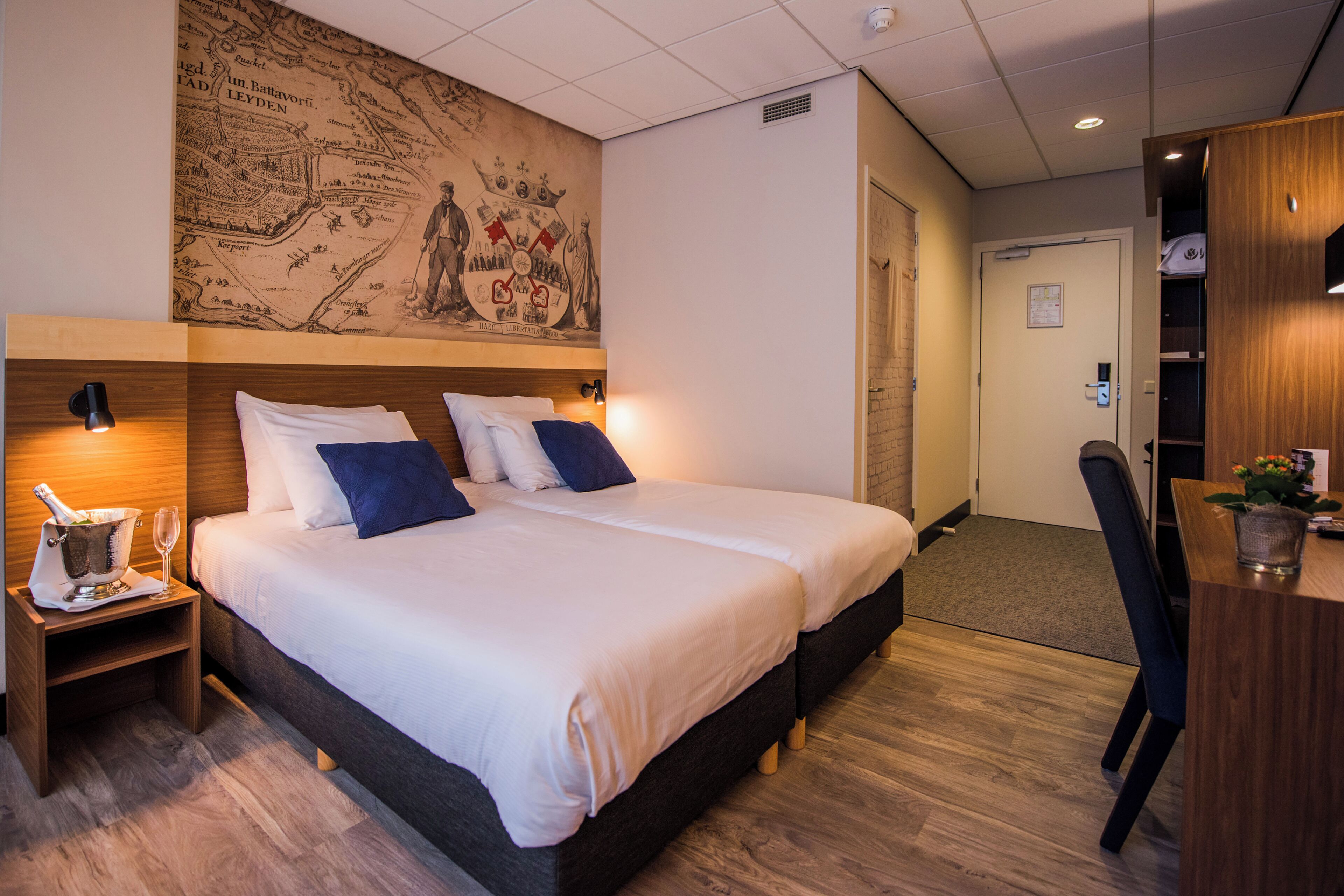 A hotel room with two beds, a wooden headboard, a map mural, a desk with a chair, and a small plant.