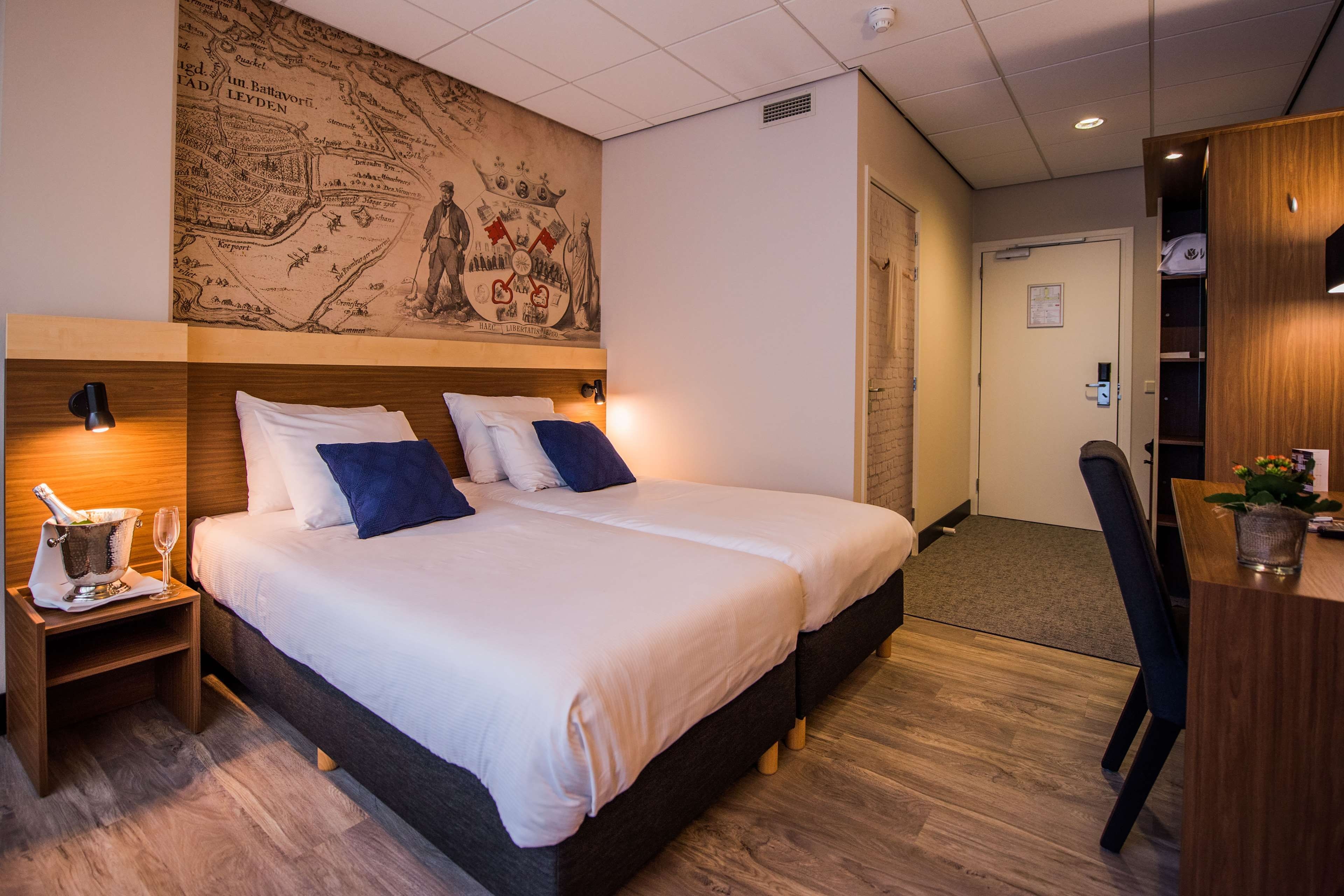 Una habitación de hotel con un gran mapa histórico en la pared, una cama con ropa blanca y almohadas azules, una mesita de noche con una botella y vasos, y un escritorio con una planta.