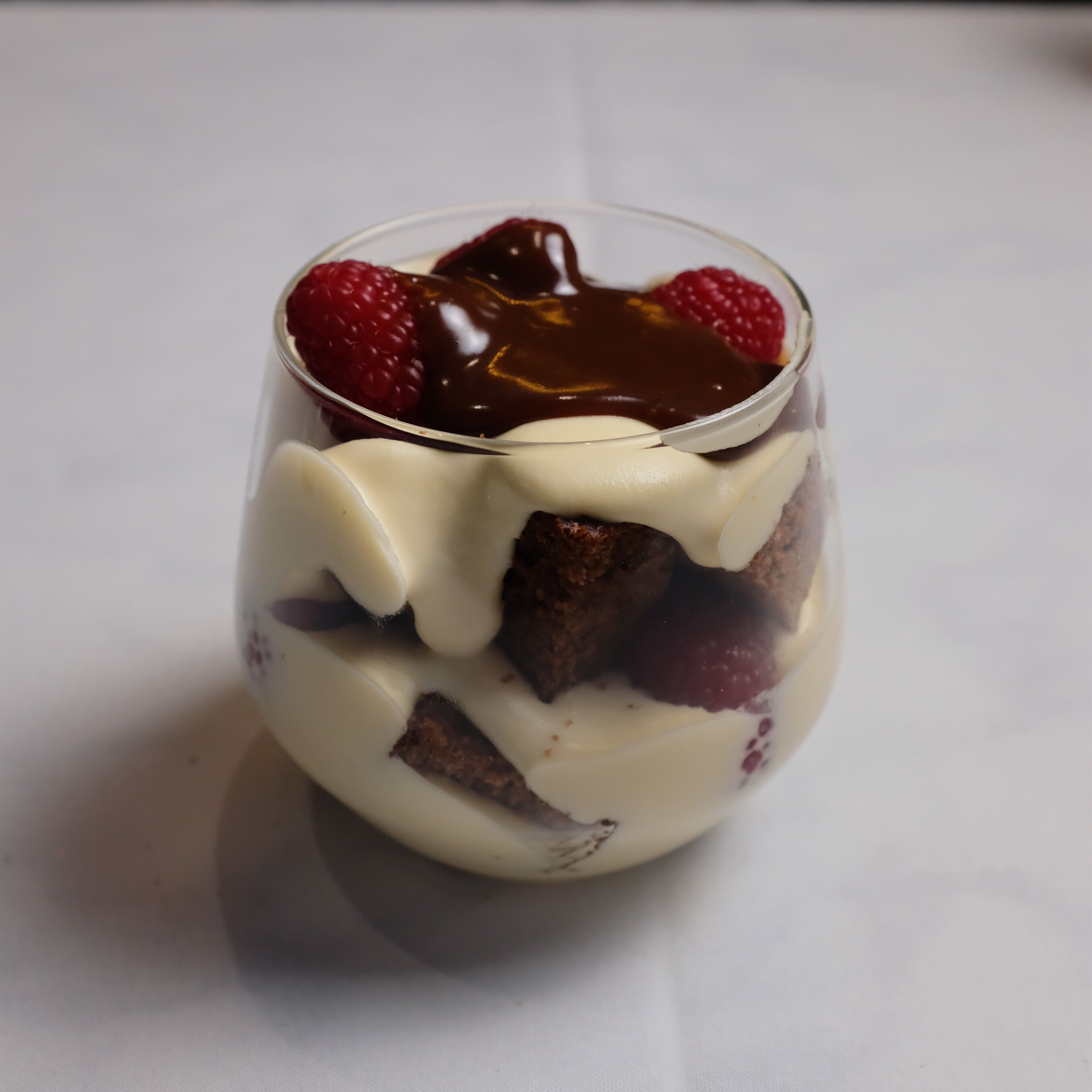 A dessert in a clear glass with layers of cream and chocolate, topped with raspberries.