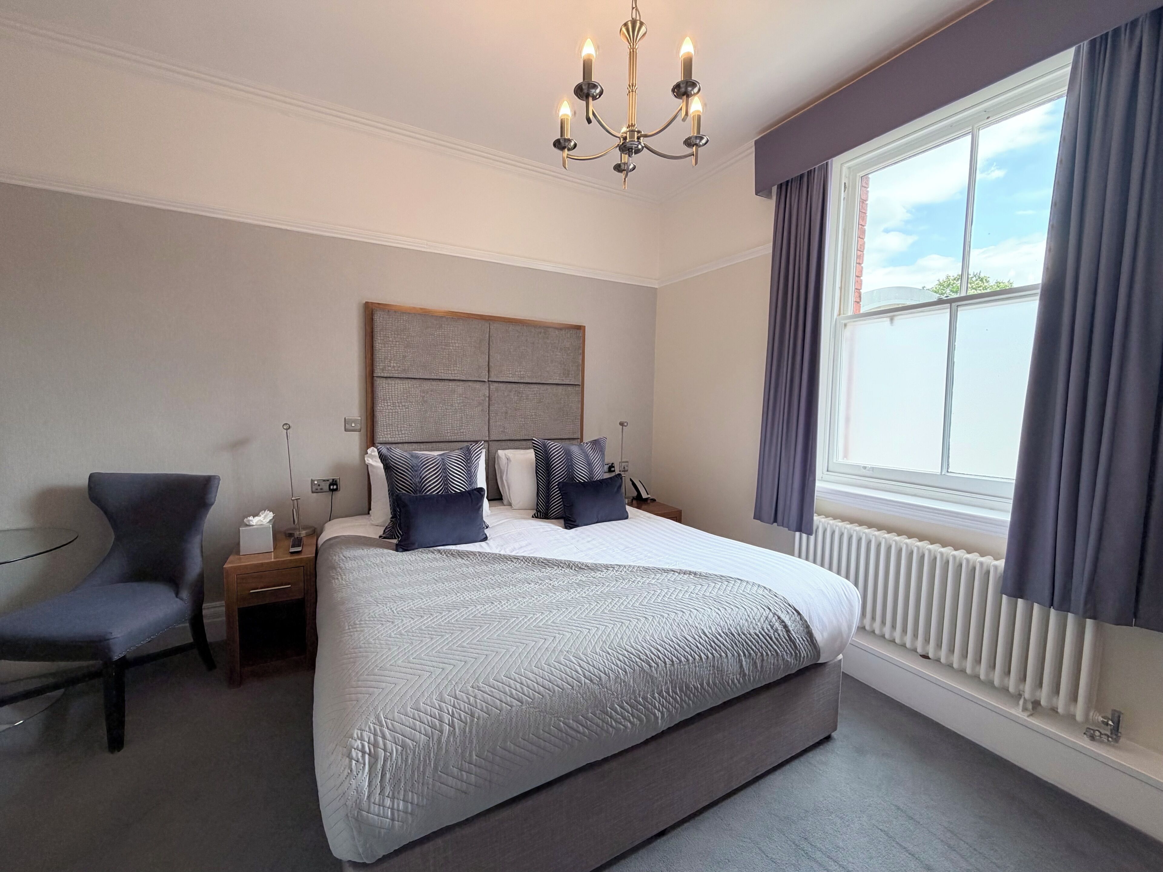 A hotel room with a large bed, a chair, a bedside table, a nightstand, a radiator, and a window with curtains.