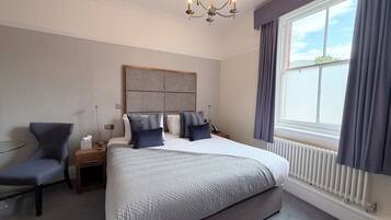 A hotel room with a large bed, a chair, a bedside table, a nightstand, a radiator, and a window with curtains.