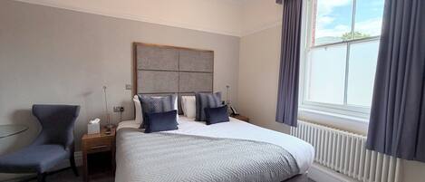 A hotel room with a large bed, a chair, a bedside table, a nightstand, a radiator, and a window with curtains.