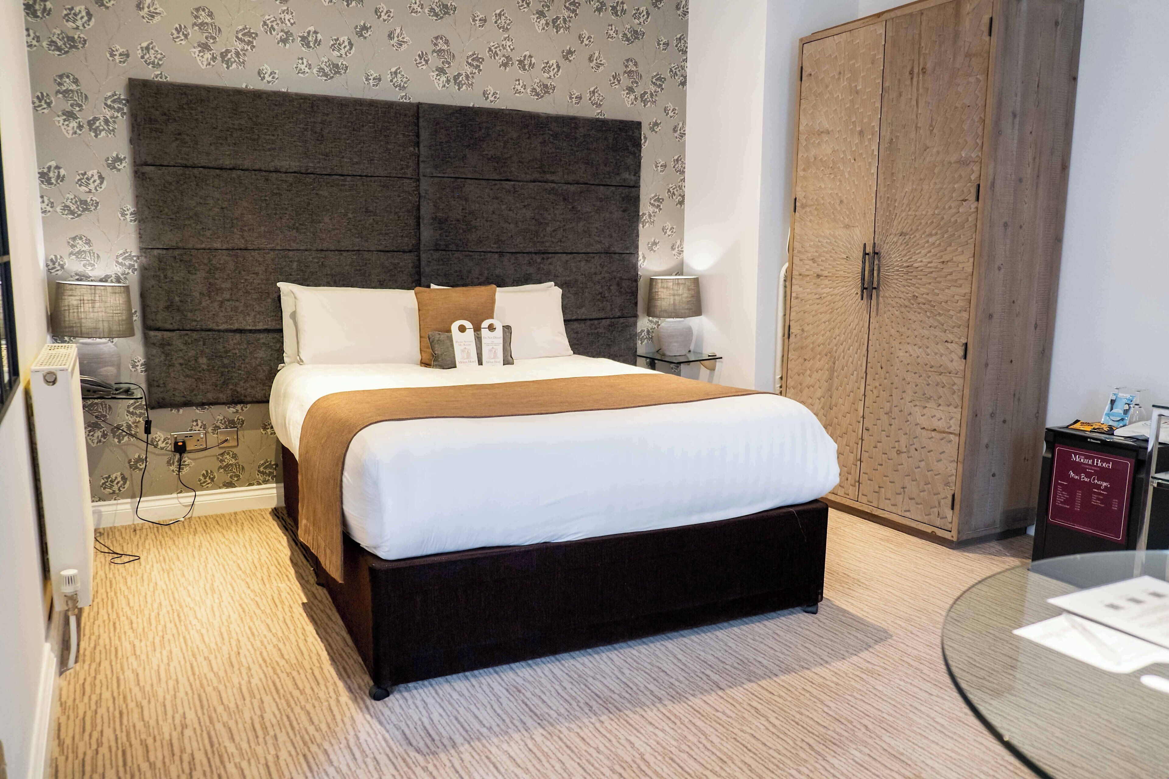 Ein Hotelzimmer mit einem großen Bett, einem dunklen Kopfteil, zwei Nachttischlampen, einem Holzschrank und einem Glastisch mit einer Speisekarte.