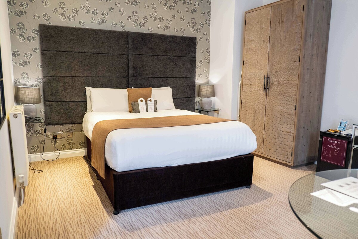 A hotel room with a large bed, a dark headboard, two bedside lamps, a wooden wardrobe, and a glass table with a menu.