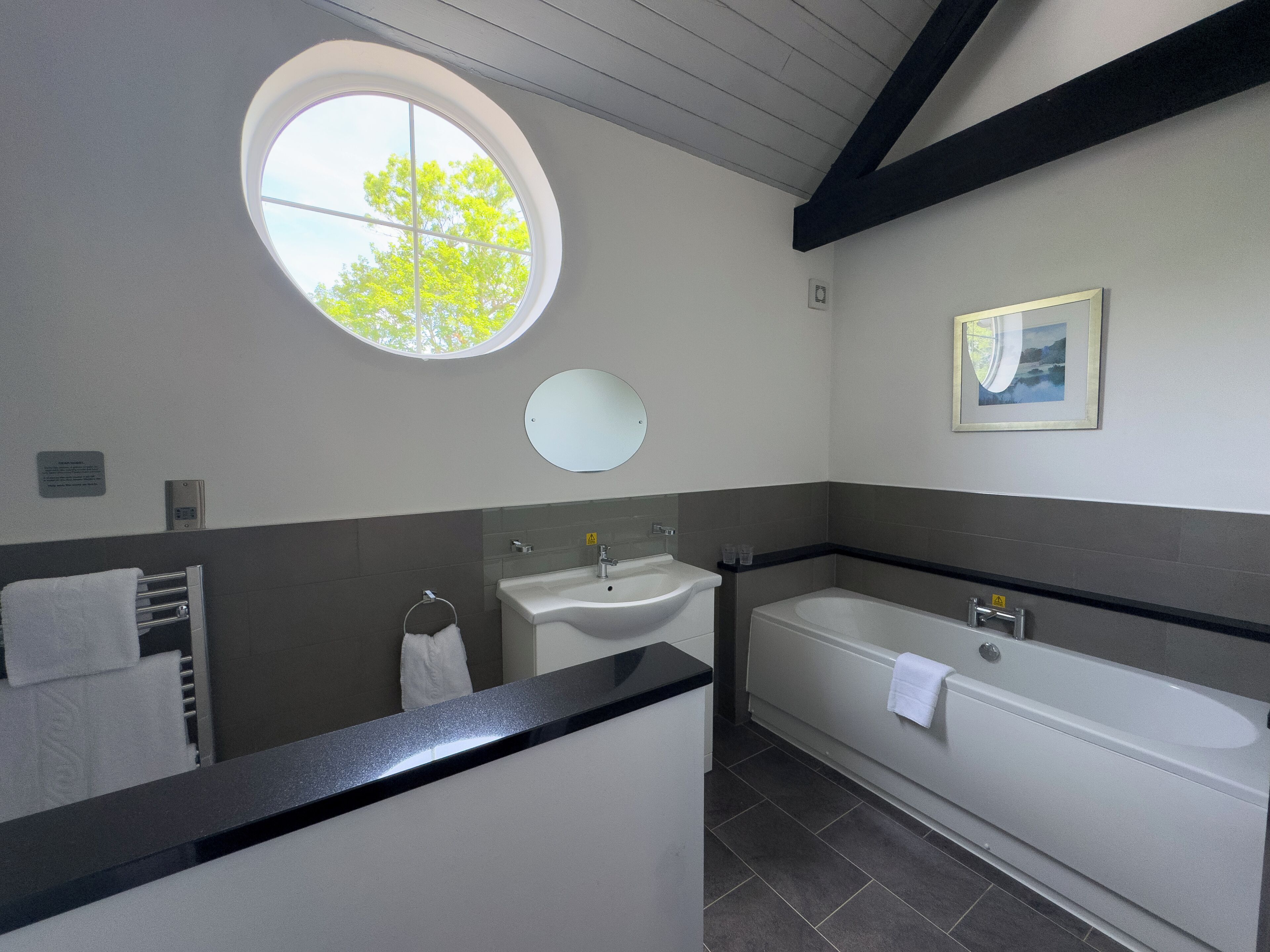 A bathroom with a round window, a sink, a mirror, a bathtub, and a towel rack.