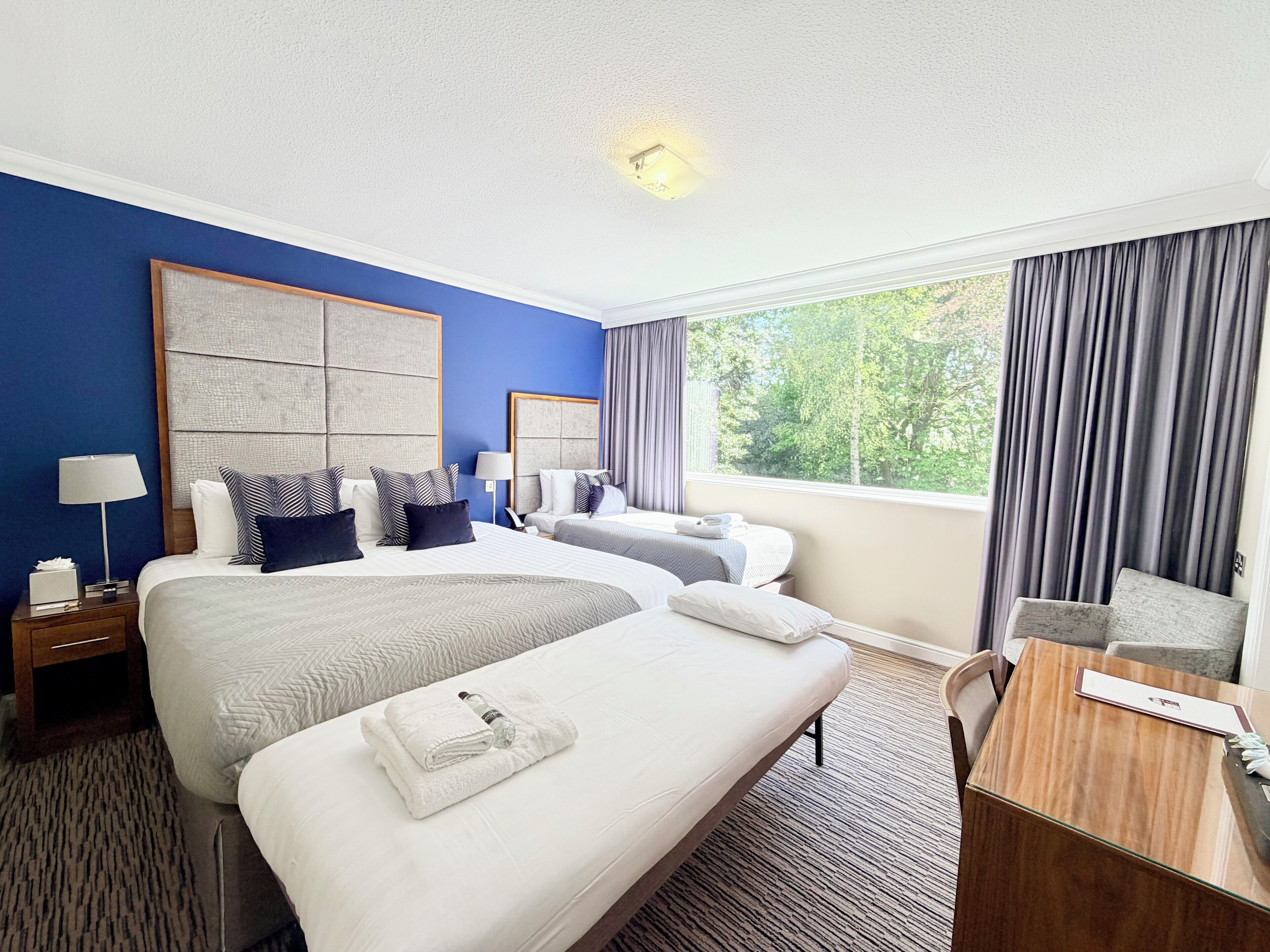 A hotel room with two beds, a desk, and a view of trees.