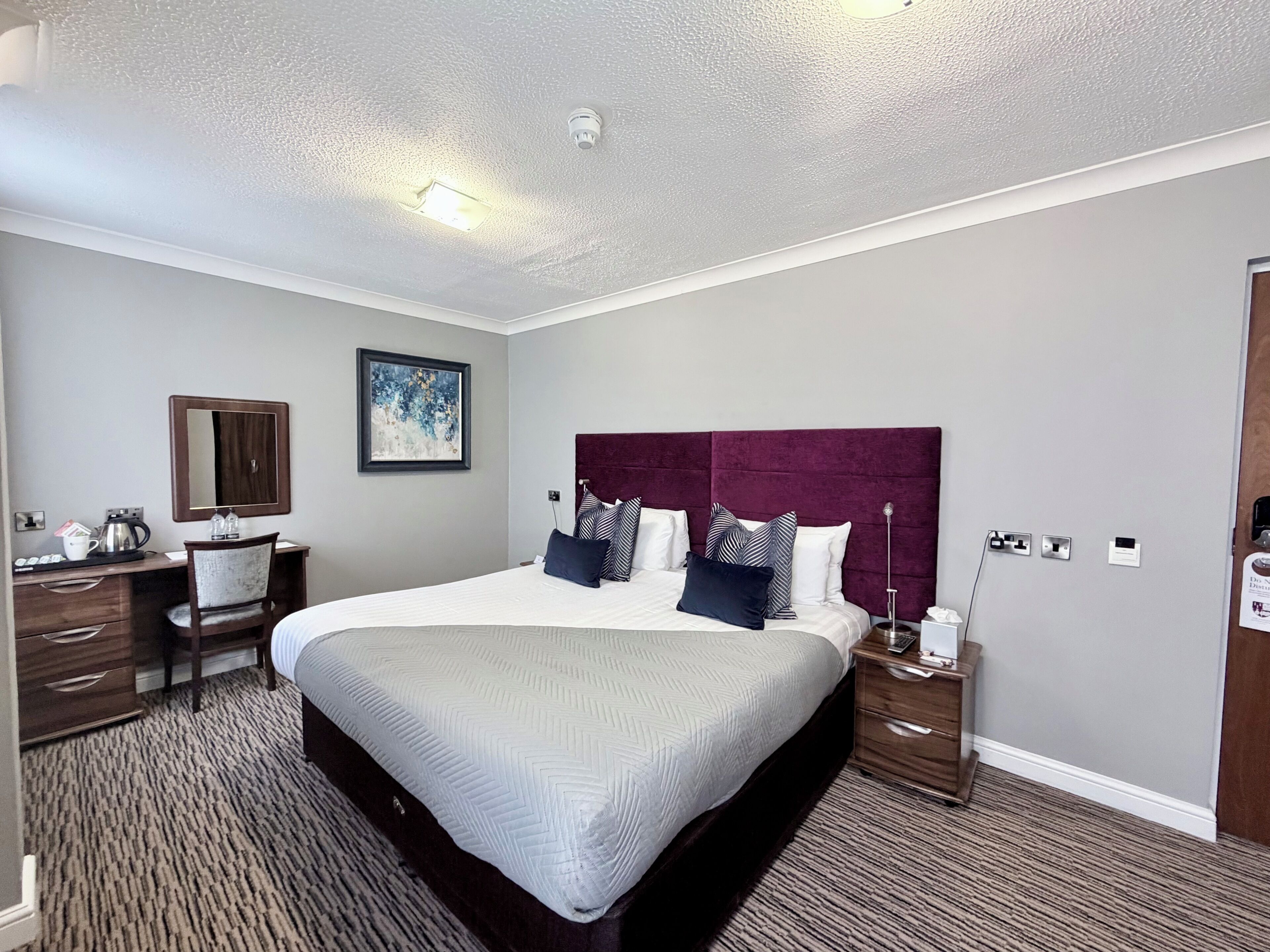 A hotel room with a large bed, a desk with a chair, a bedside table with a lamp, and a wall-mounted mirror.