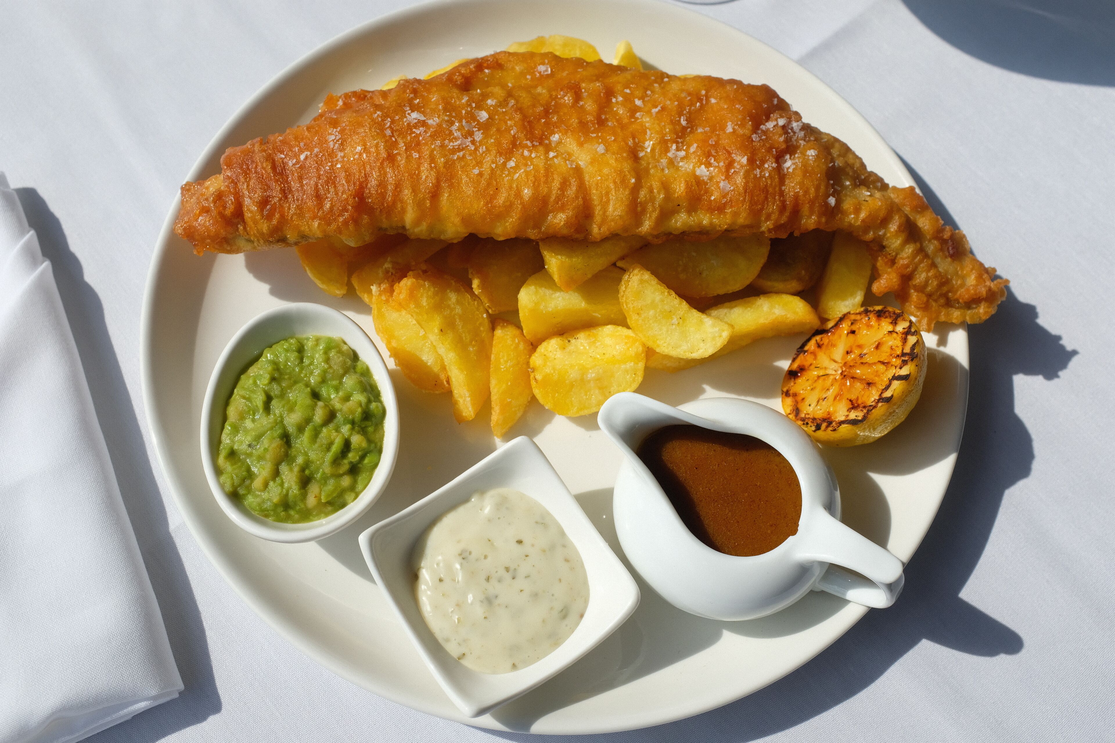 Ein Teller mit Fish and Chips, bestehend aus einem großen panierten Fischfilet, goldbraunen Pommes Frites, einer kleinen Schale Erbsenpüree und jeweils einer kleinen Schale Tartarsauce und Bratensoße.