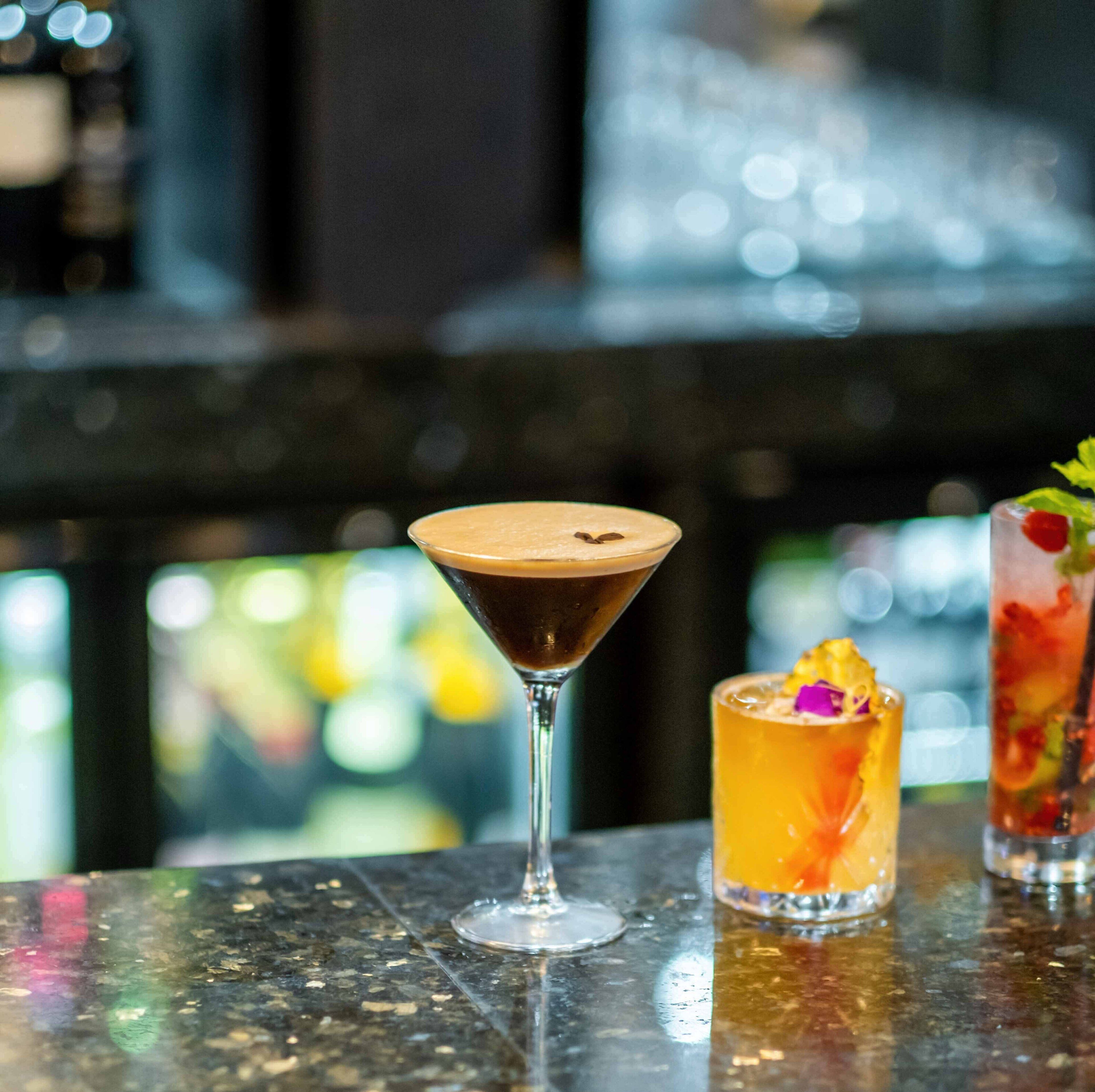 Una barra con tres bebidas: un martini, un cóctel con adorno de fruta y una bebida colorida con pajita.