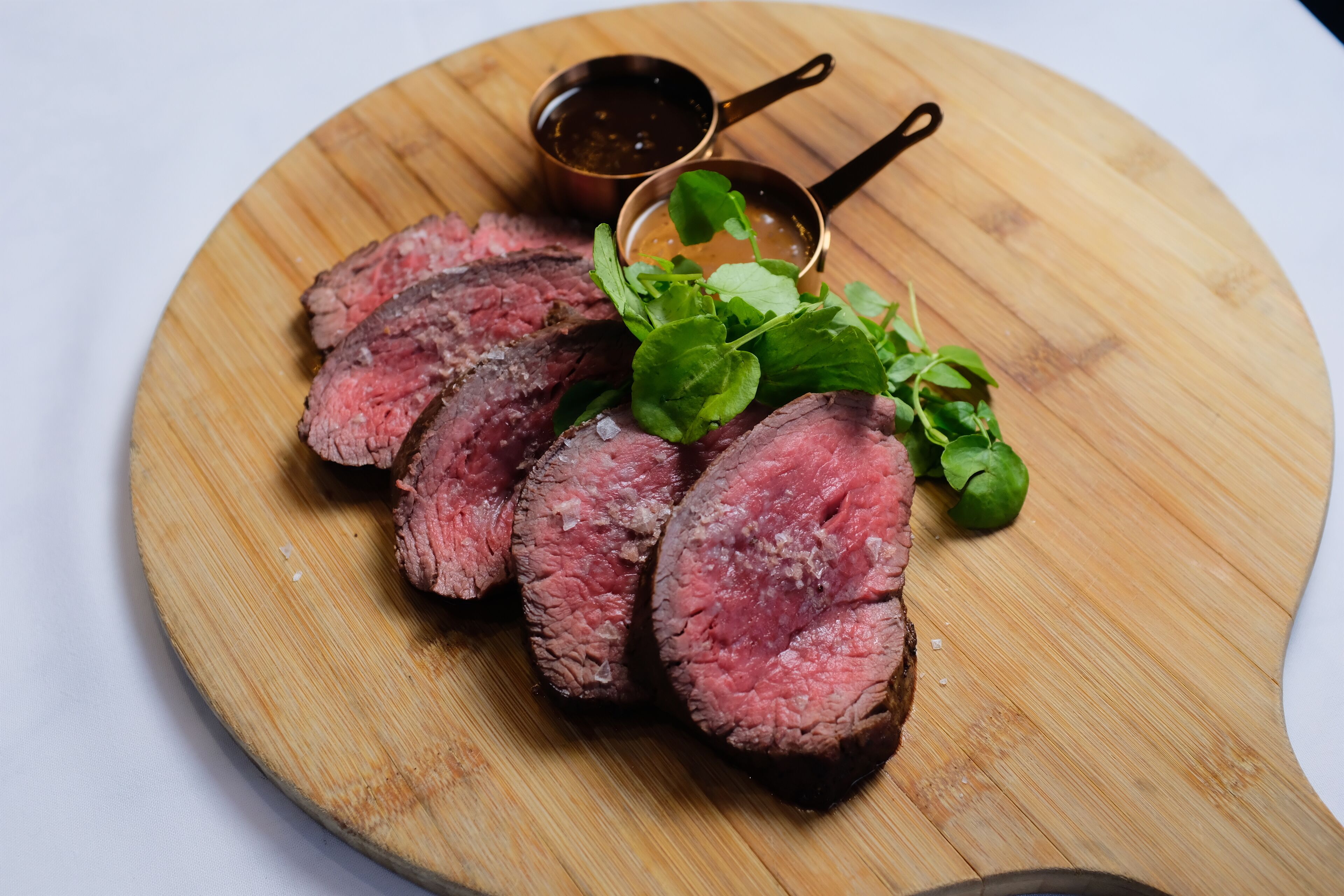 A wooden cutting board with sliced steak, greens, and two small bowls of sauce.