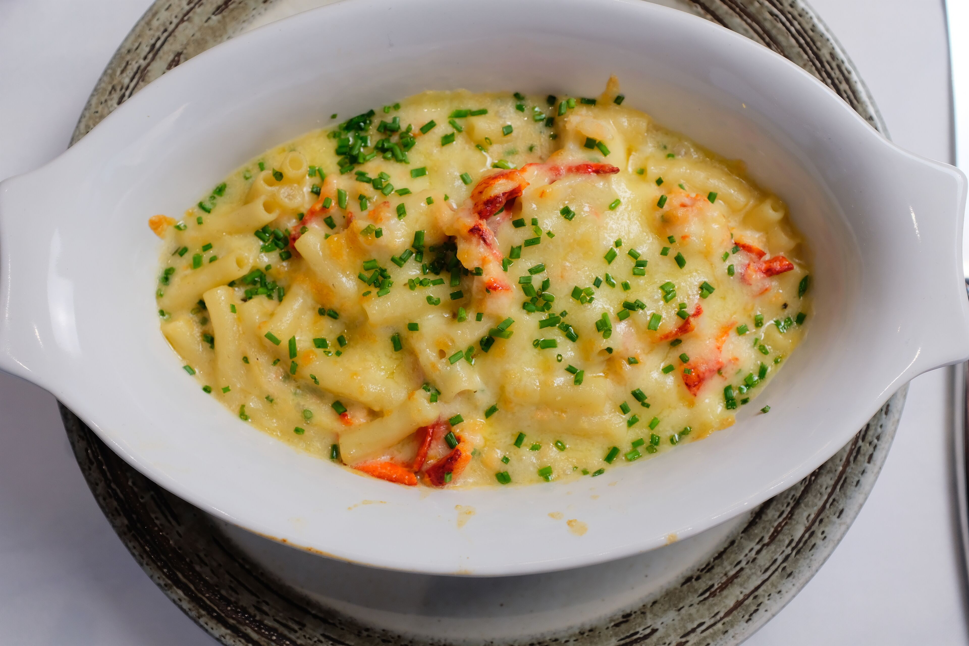 Un plato blanco ovalado con pasta, queso derretido y cebollines, sobre un posavasos tejido.