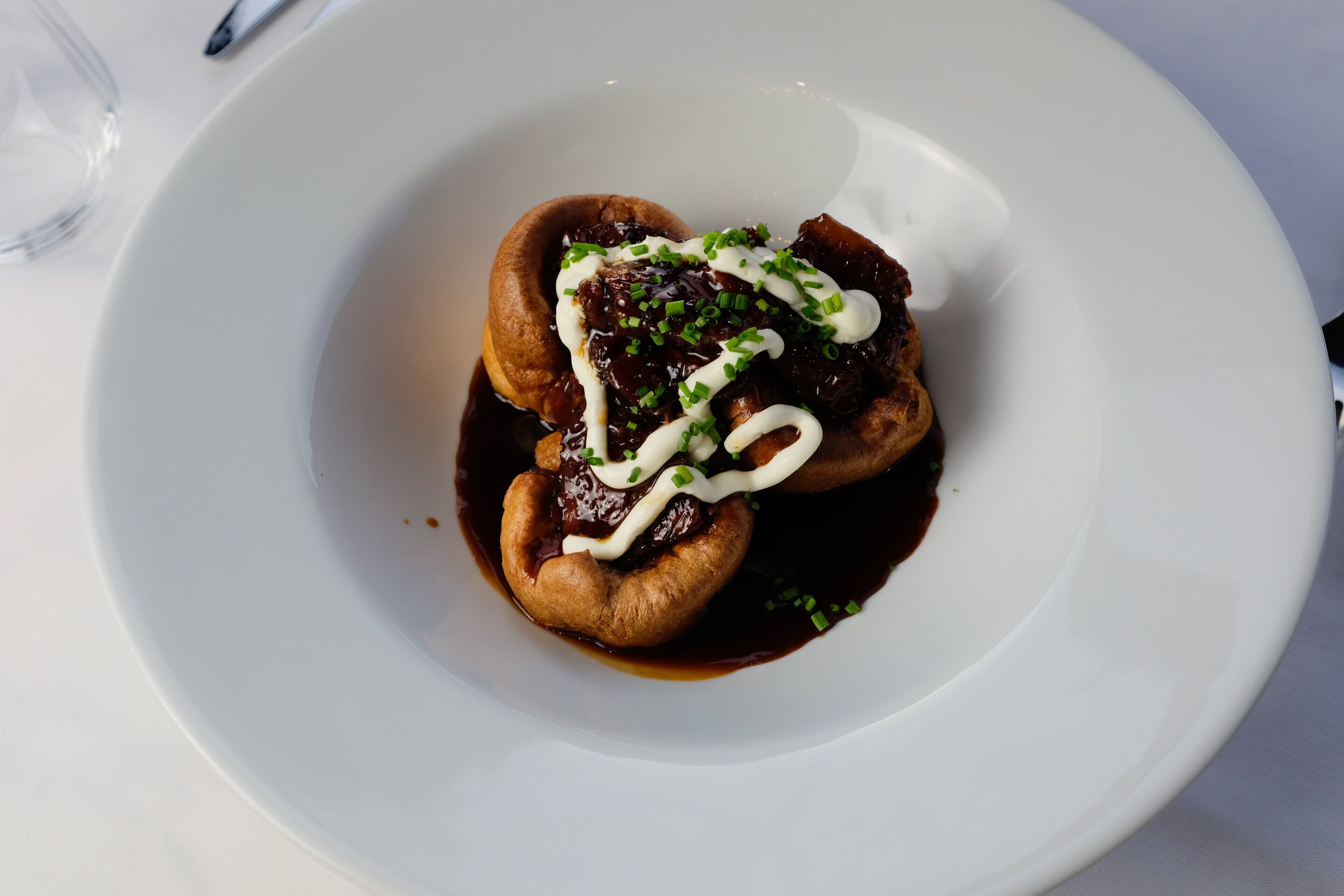 A white plate with a dessert consisting of a pastry, sauce, and a dollop of cream, garnished with green herbs.