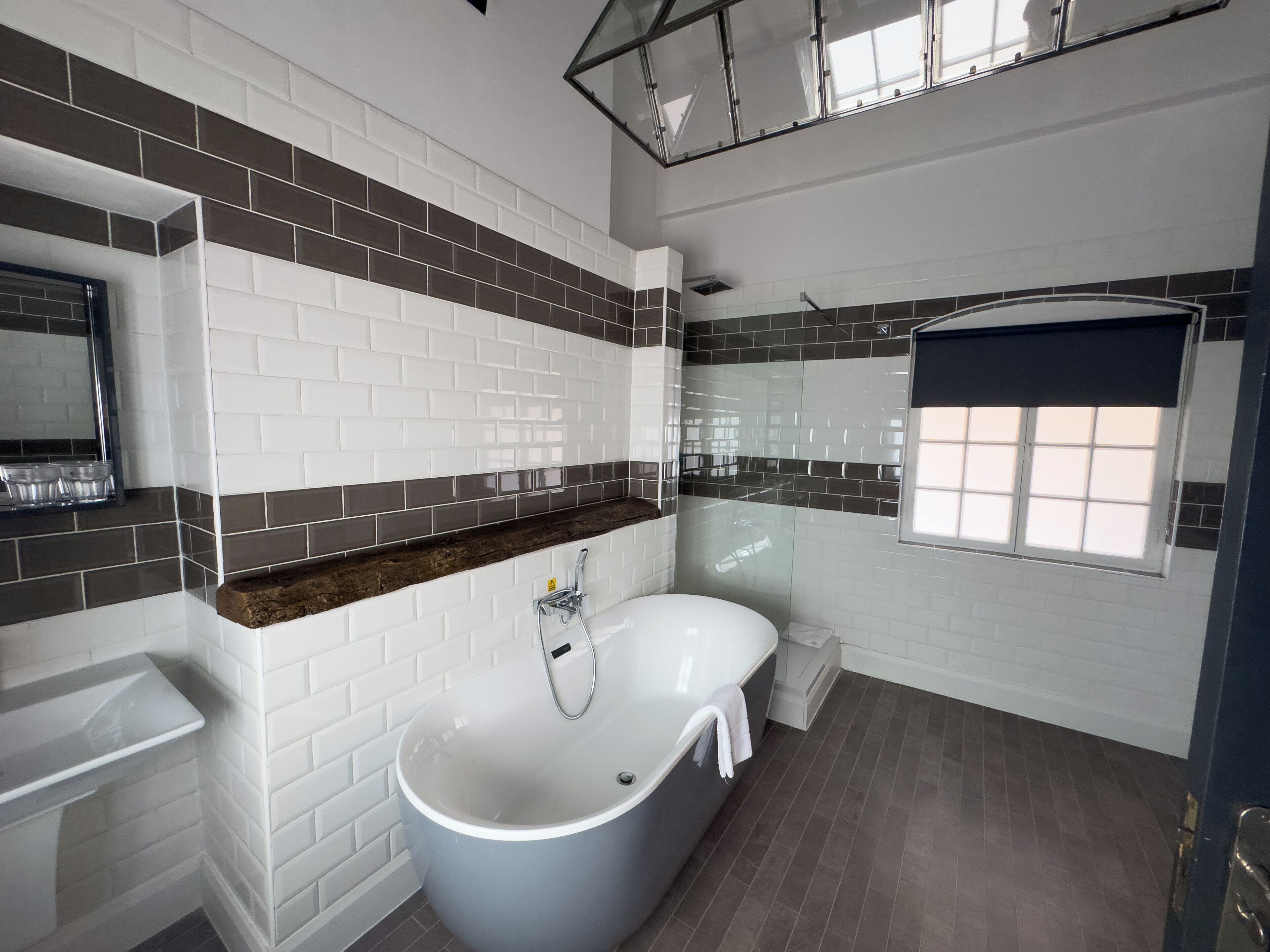 Un baño moderno con una bañera independiente, baldosas blancas y oscuras, una ventana con vistas y un espejo sobre el lavabo.