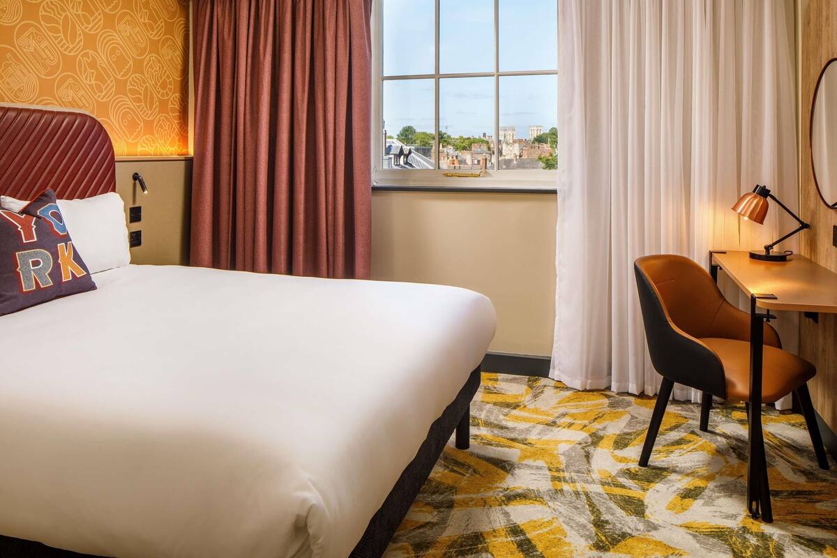 A hotel room with a large bed, a desk with a chair, a window with a view of buildings, and a patterned carpet.