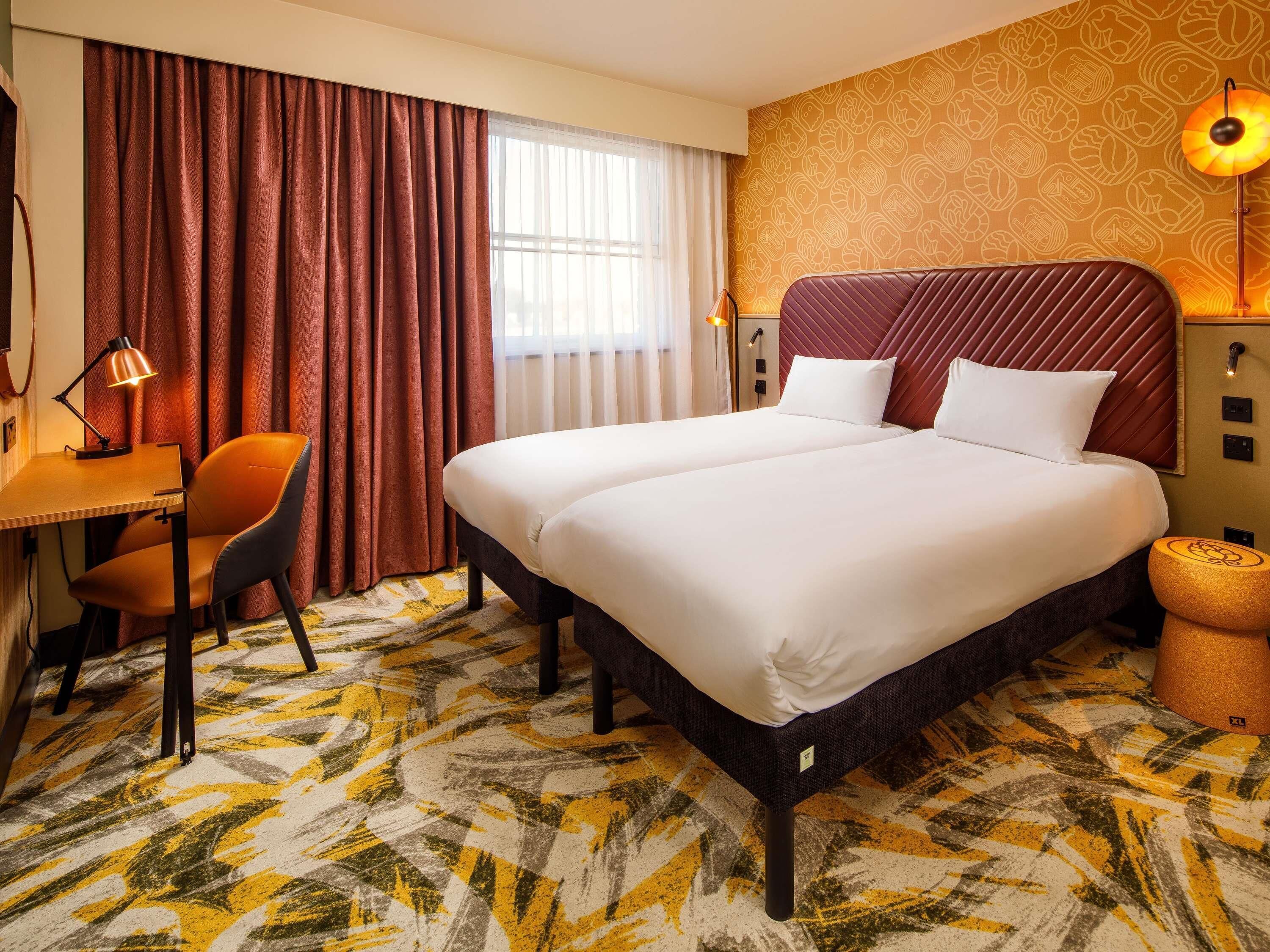 A hotel room with a large bed, a desk with a chair, a lamp, and a patterned carpet.