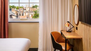 Quarto de hotel com cama, escrivaninha com luminária, cadeira, espelho grande e vista para a paisagem urbana através de uma janela com cortinas.