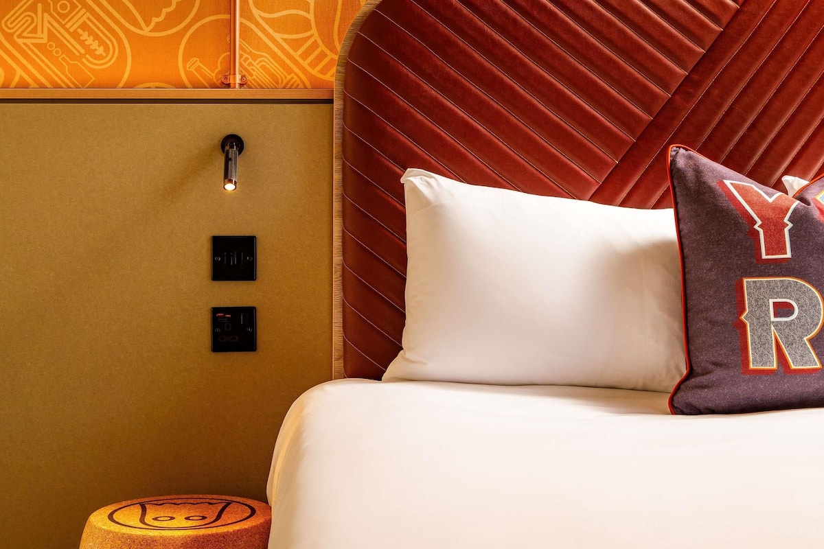 Habitación de hotel con cama, cabecera, mesita de noche y panel de control en la pared.
