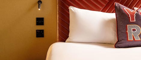 Habitación de hotel con cama, cabecera, mesita de noche y panel de control en la pared.