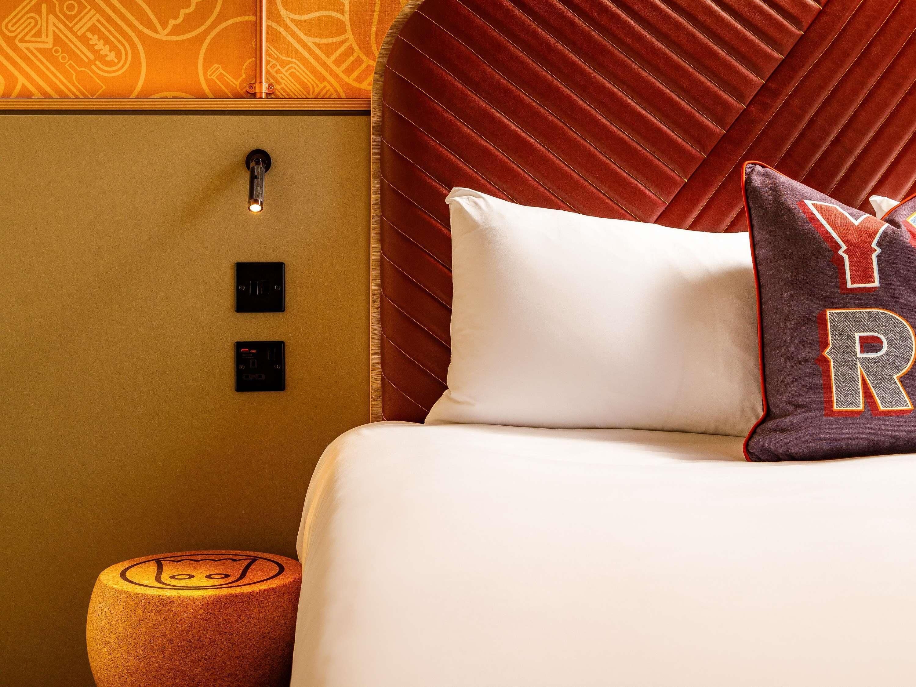 A hotel room with a bed, a headboard, a nightstand, and a wall-mounted control panel.