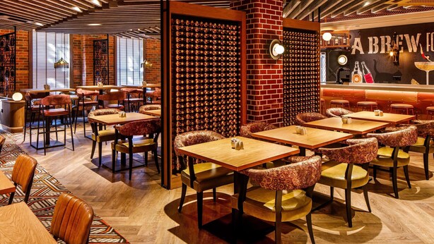 Interior de restaurante com vigas de madeira no teto, paredes de tijolo e adega. Mesas e cadeiras dispostas para refeições.