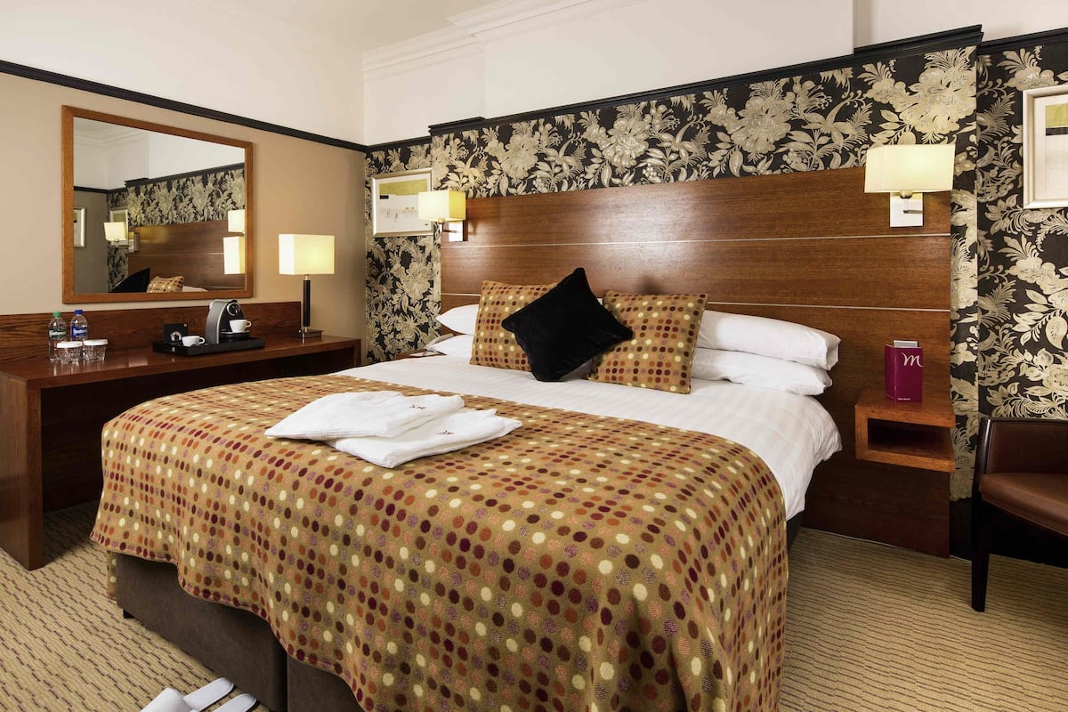 Una habitación de hotel con una cama grande, cabecera de madera, mesita de noche, escritorio con lámpara, espejo y una silla.