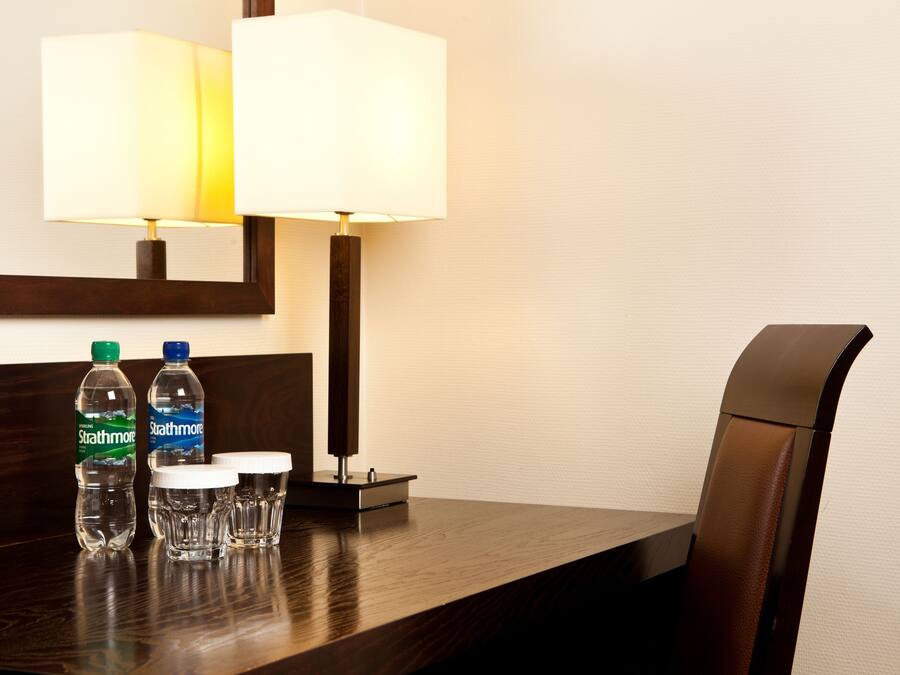 Quarto de hotel com cabeceira de madeira, duas luminárias, duas garrafas de água e dois copos para shots sobre uma mesa de madeira.