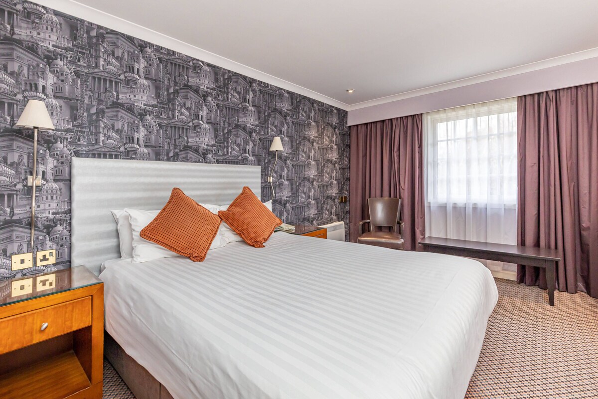 Een hotelkamer met een groot bed, twee kussens, een nachtkastje, een lamp, een stoel, een raam met gordijnen en behang met een patroon.