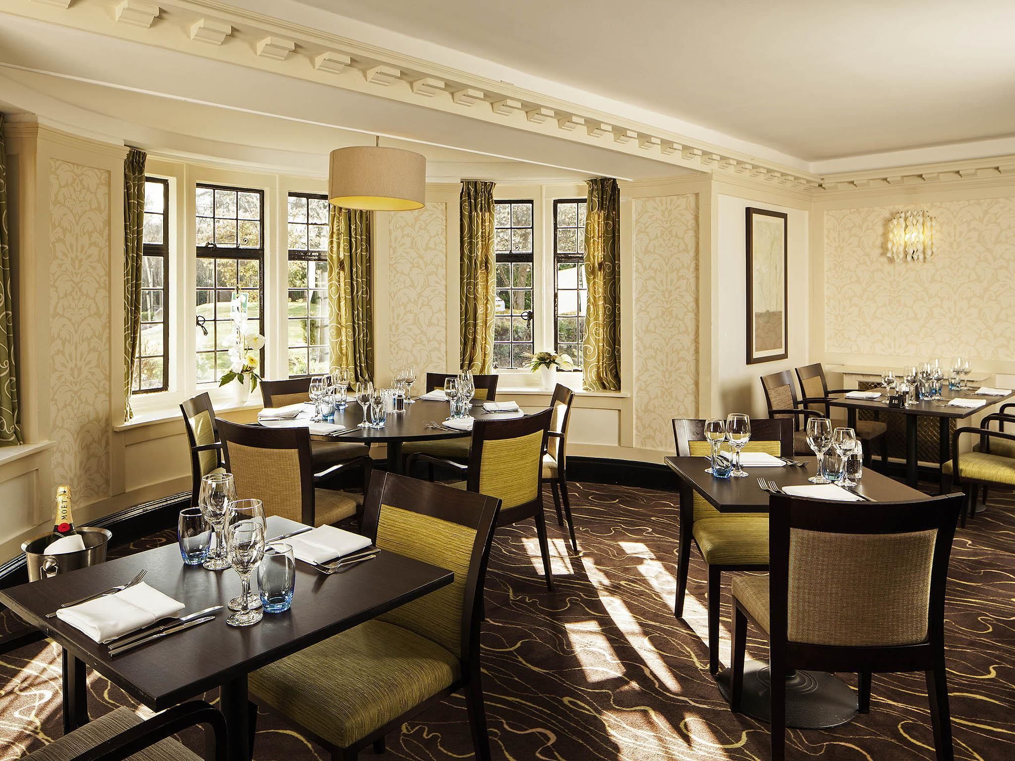 A formal dining room with multiple tables set for service, large windows with curtains, and a patterned carpet.