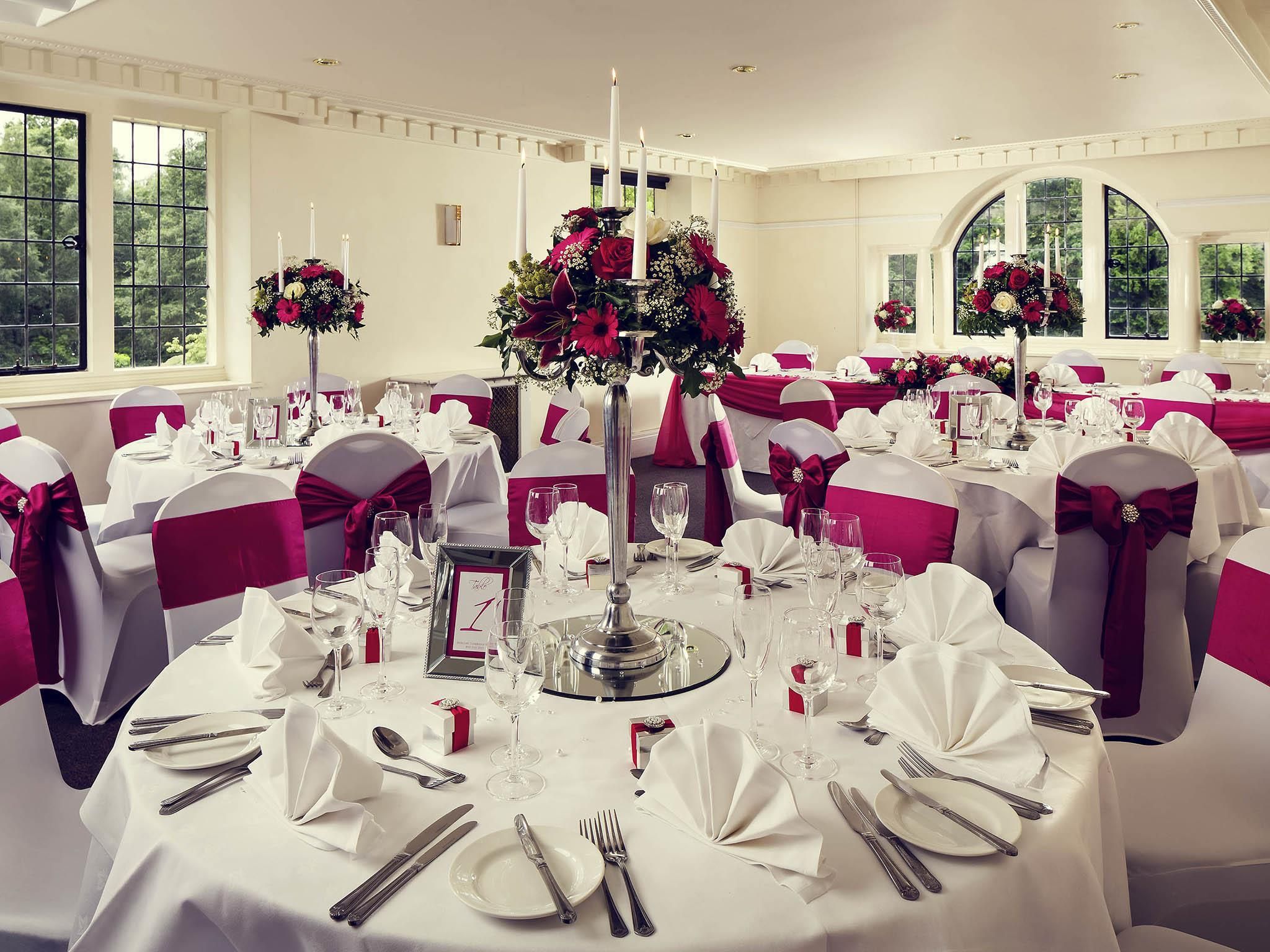 A banquet hall with round tables set for a formal event, featuring white tablecloths, red sashes, and floral centerpieces.