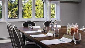 Un salón de conferencias con una gran mesa, sillas, un ventanal con vistas a árboles y varios objetos como vasos, papeles y recipientes.