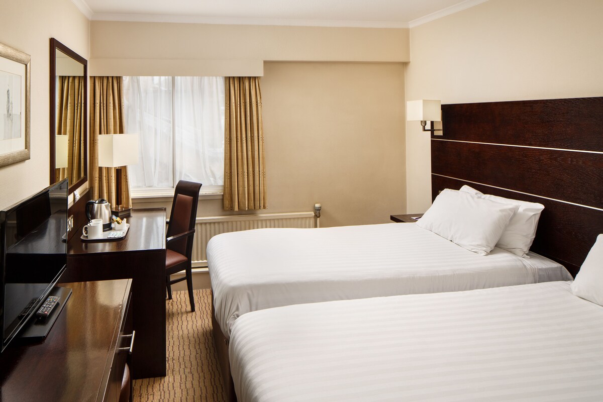 Een hotelkamer met twee bedden, een bureau met een laptop, een stoel, een lamp en een raam met gordijnen.