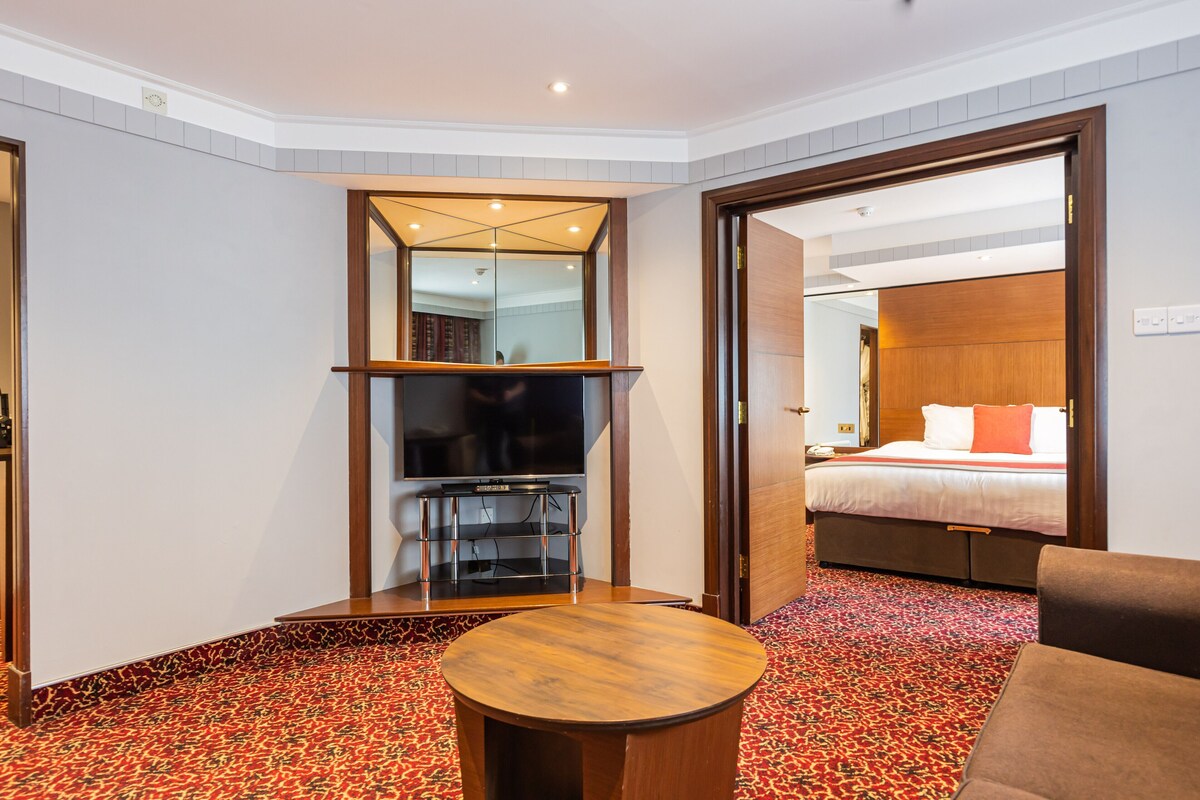 Een hotelkamer met een groot bed, een televisie, een houten bijzettafeltje en een kamerbreed tapijt met een patroon.
