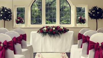 A wedding ceremony setup with white chairs, a floral centerpiece, and a large arched window with greenery outside.