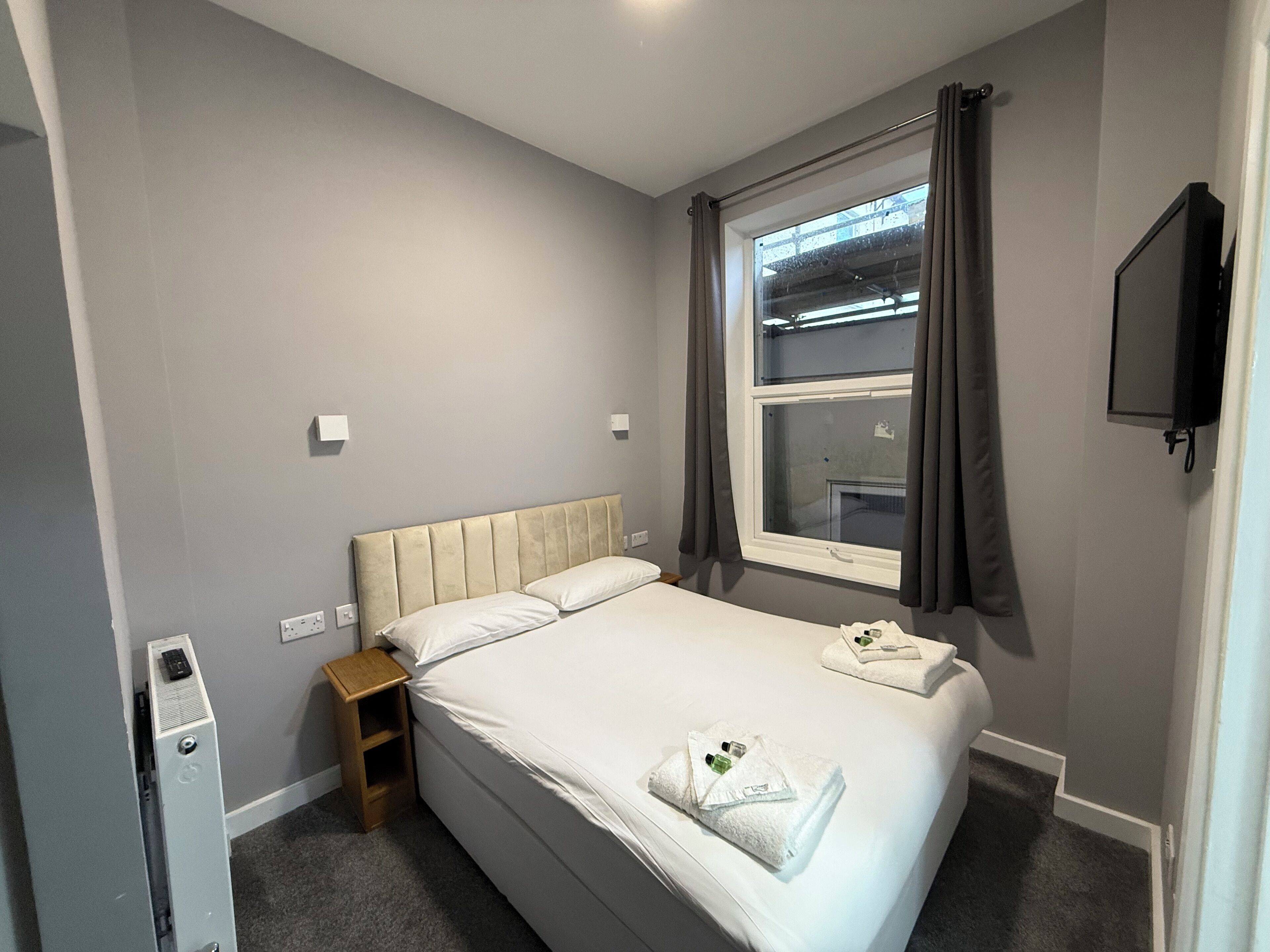 A neatly made bed with white linens and two towels, a bedside table with a lamp, a window with curtains, and a TV mounted on the wall.