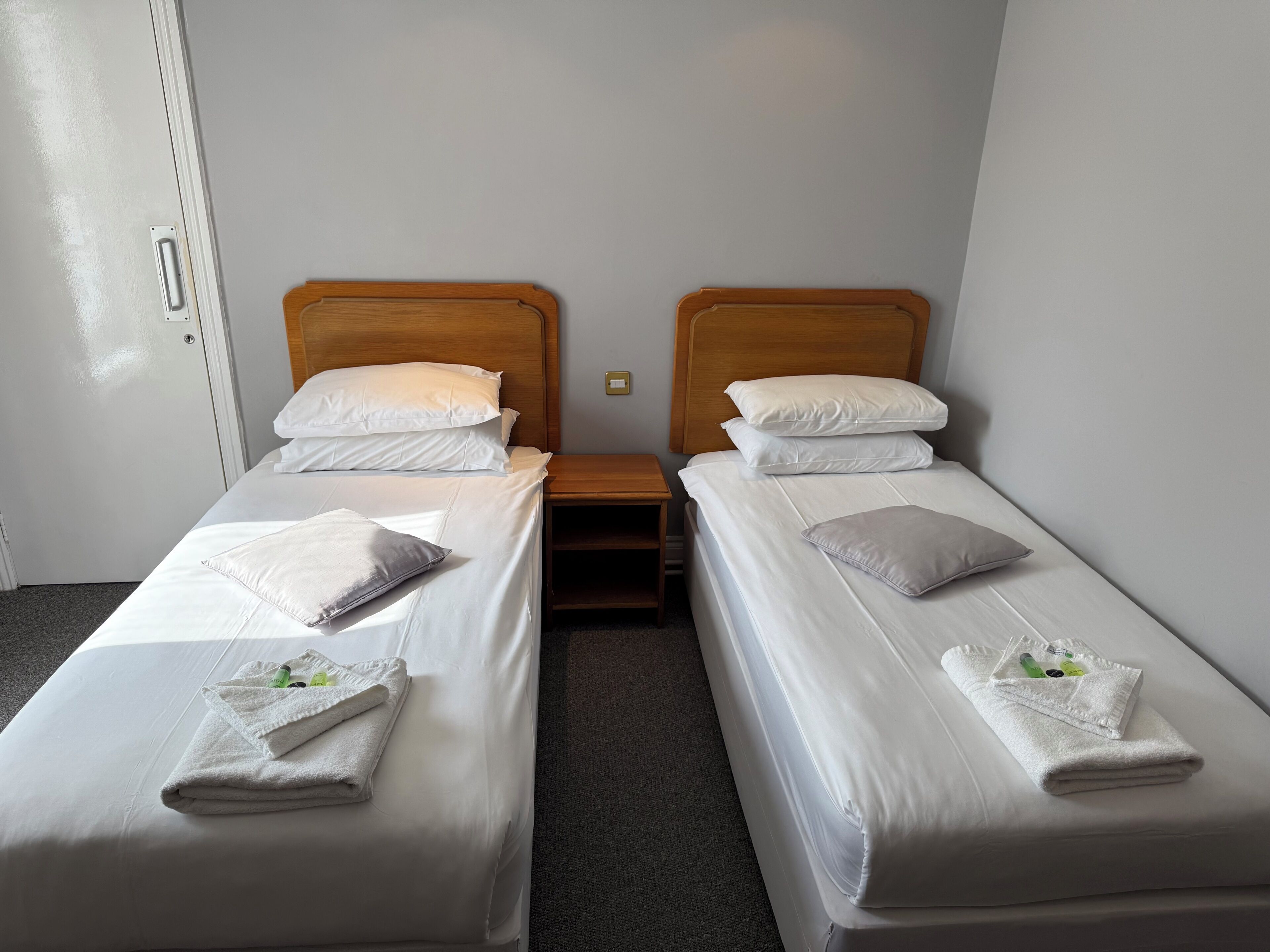 Habitación de hotel con dos camas individuales, cada una con sábanas y almohadas blancas, cabeceros de madera y una pequeña mesita de noche entre ellas.