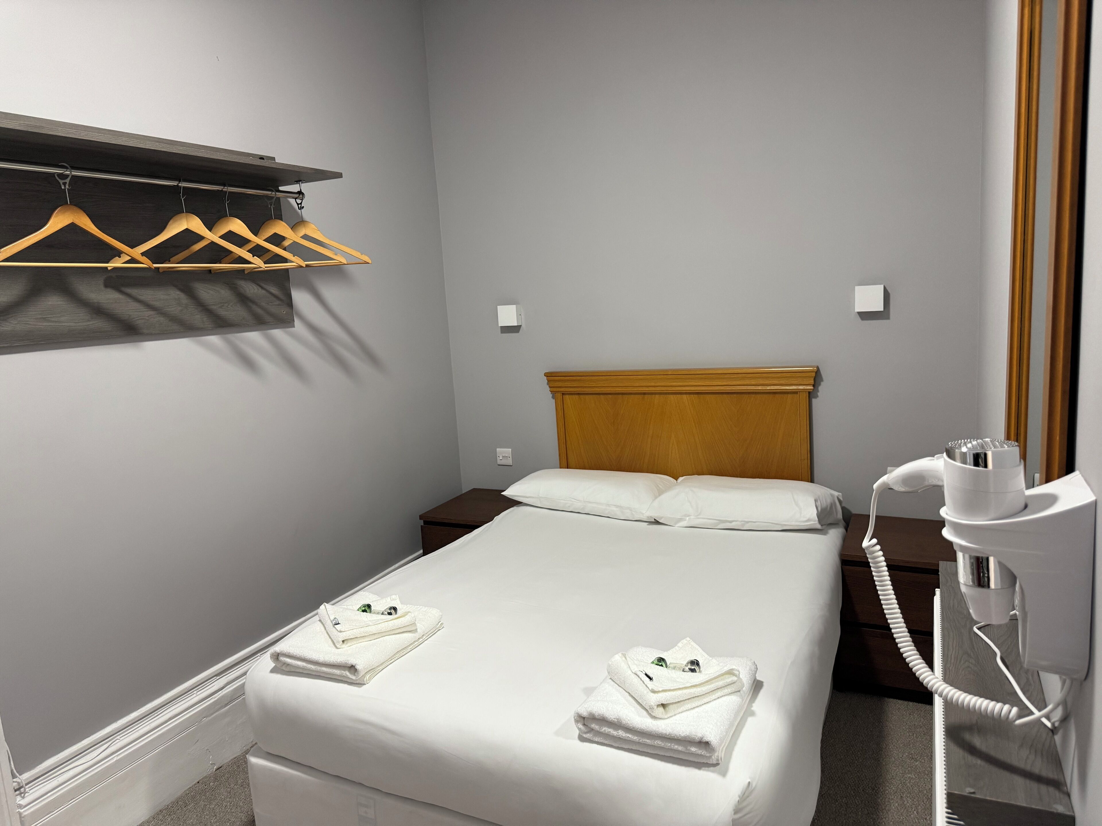 Ein Hotelzimmer mit Bett, Holz-Kopfteil, Nachttisch, Fön und Kleiderstange mit Kleiderbügeln.