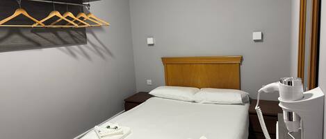 Ein Hotelzimmer mit Bett, Holz-Kopfteil, Nachttisch, Fön und Kleiderstange mit Kleiderbügeln.
