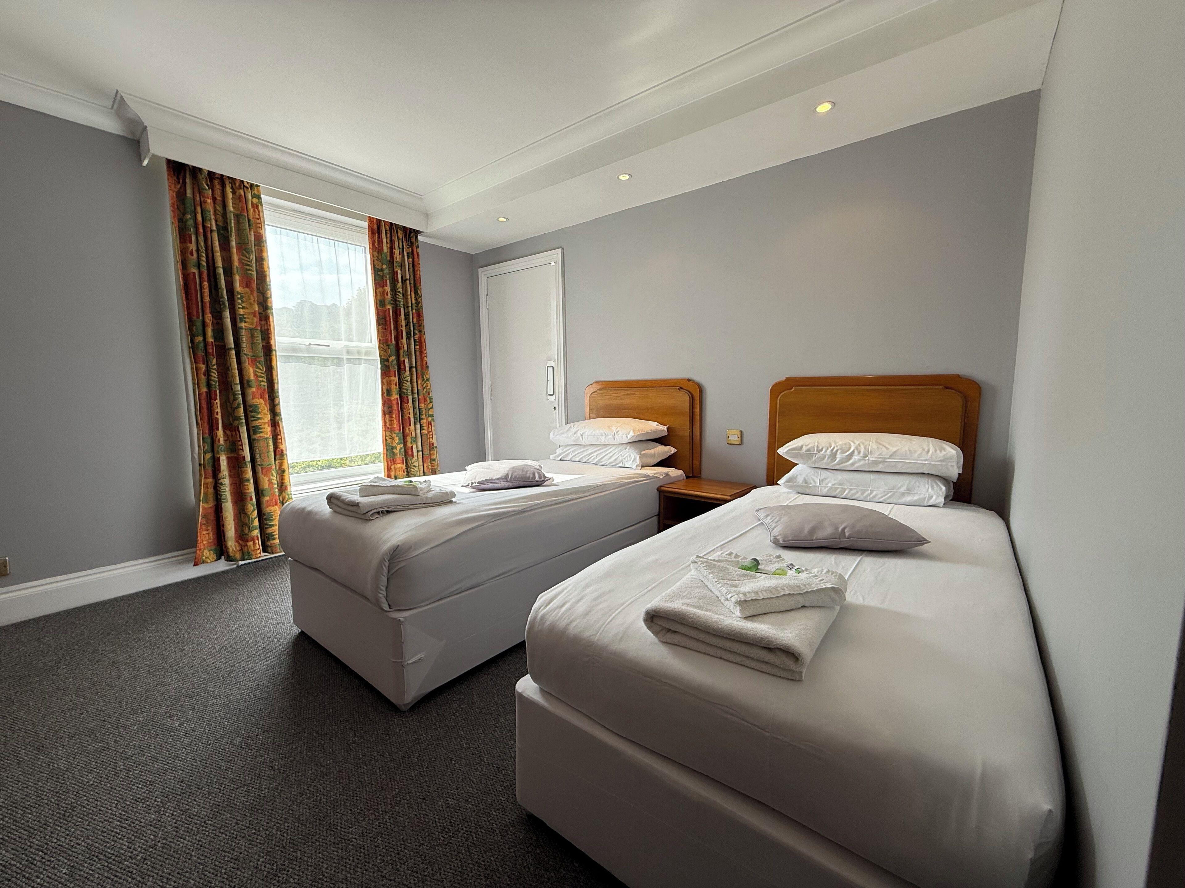 Habitación de hotel con dos camas, una ventana con cortinas, una mesita de noche y piso alfombrado.