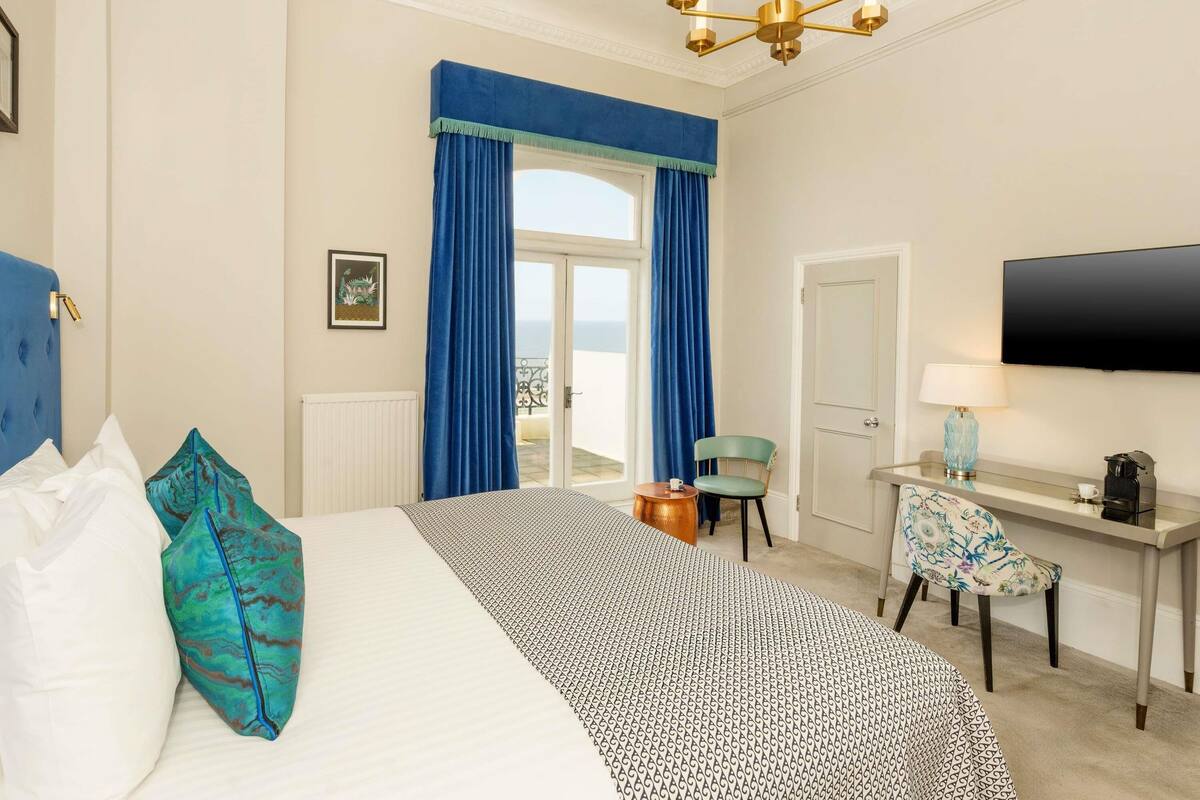 A hotel room with a bed, a desk, a chair, a television, and a view of the sea.