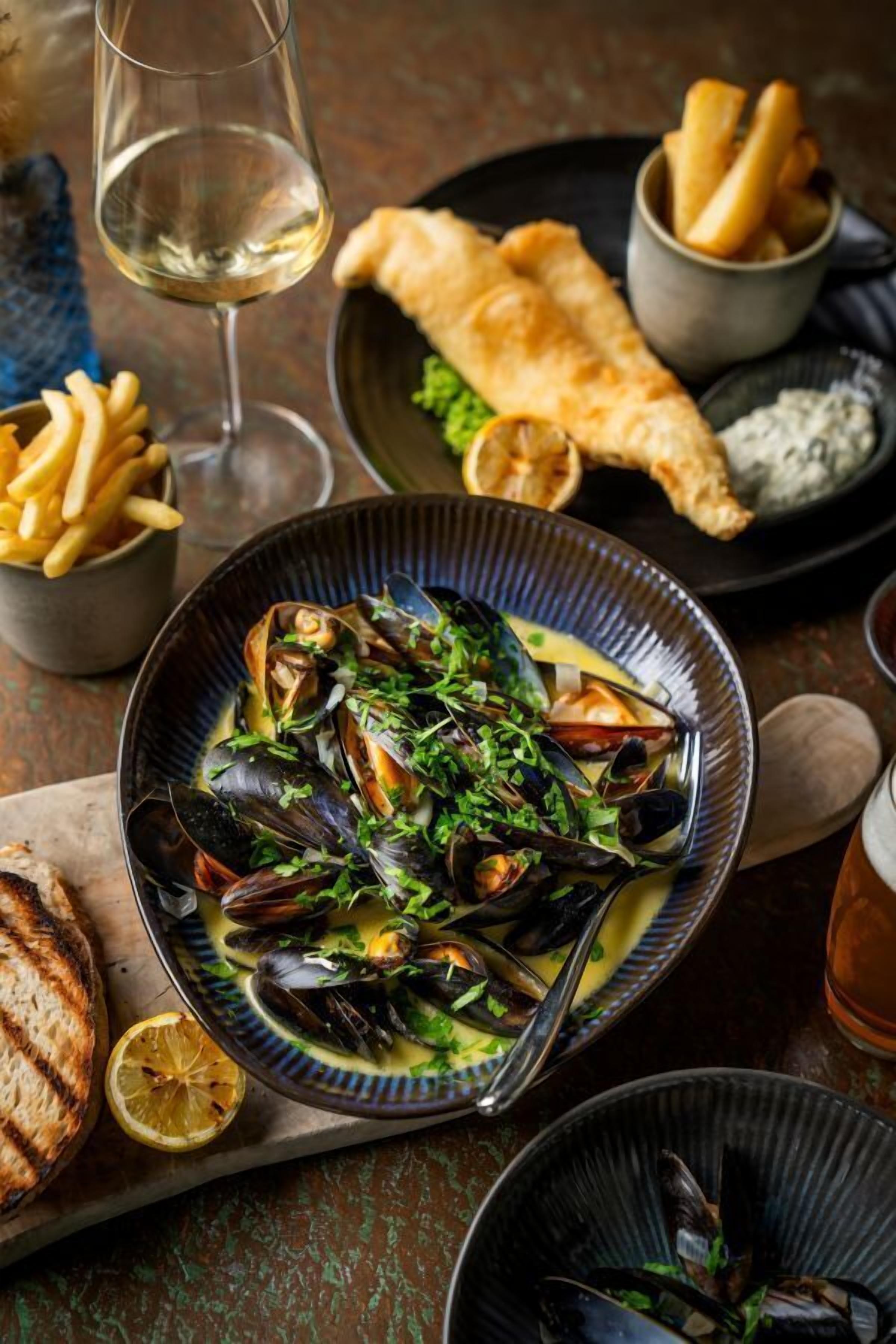 Un bol de mejillones con una rodaja de limón, una copa de vino blanco y un plato de papas fritas.
