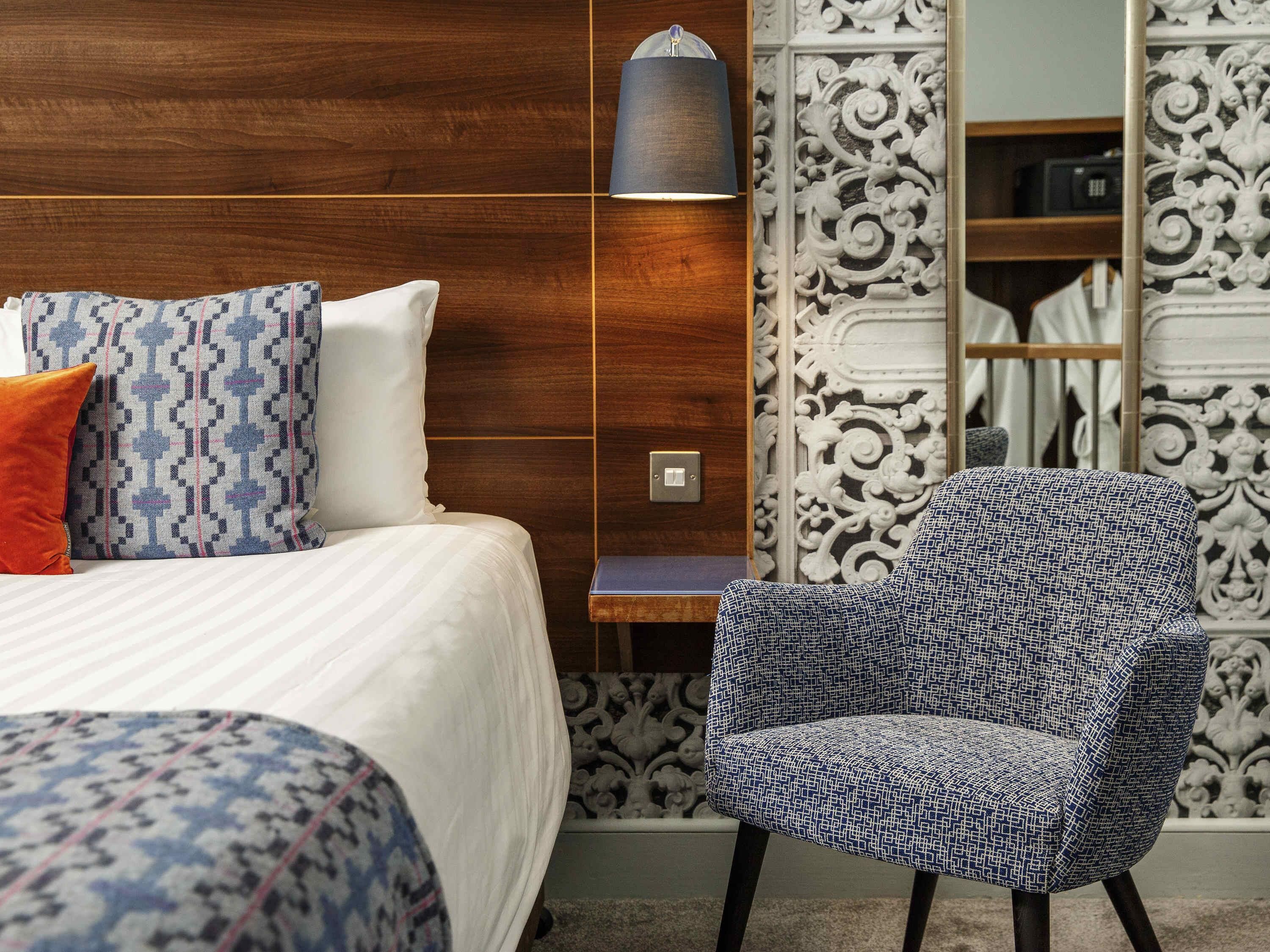 A hotel room with a bed, a patterned armchair, a nightstand, and a decorative wall panel.