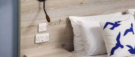 Una cama con ropa de cama blanca y un almohadón con diseños de pájaros azules, una mesita de noche, una lámpara de pared y un enchufe.