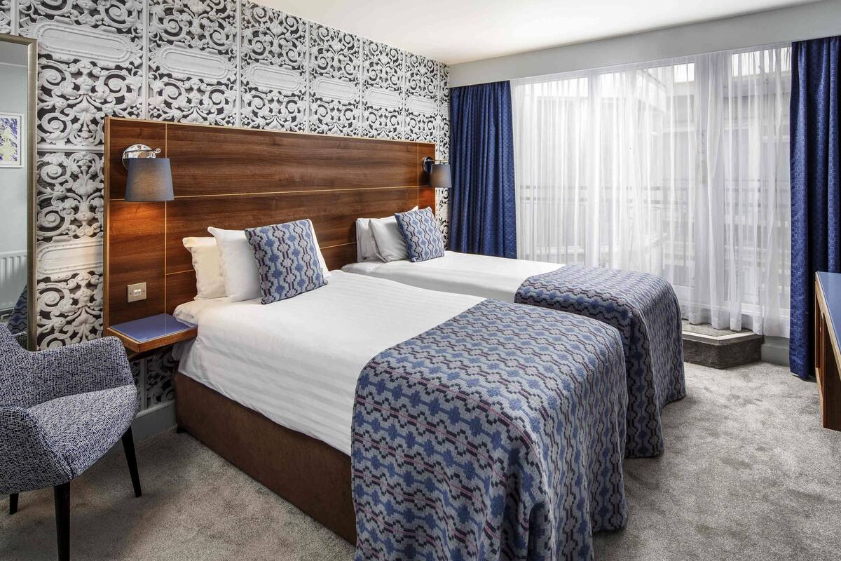 A hotel room with two beds, a wooden headboard, a nightstand, a chair, and a large window with curtains.